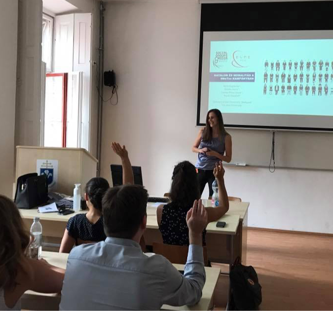 Boglarka standing in front of a whiteboard presenting to a class. Couple of students holding their hands up.