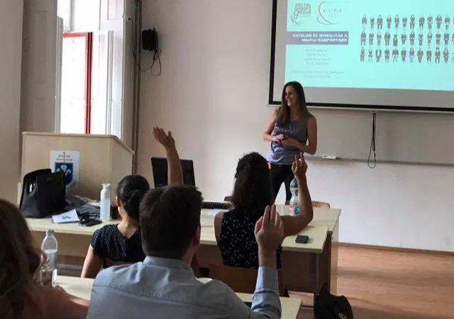 Boglarka standing in front of a whiteboard presenting to a class. Couple of students holding their hands up.
